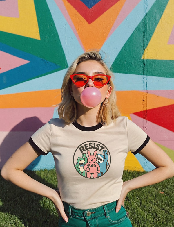 An off-white cream-colored unisex ringer t-shirt with contrasting black trim on the crewneck collar and short sleeves, laid flat on a solid background. In the center of the chest is a large circular emblem with a thick black border. Inside the circle, bold, black, sans-serif text arches across the top, spelling "RESIST." Below the text are three cartoon characters: a green frog in a suit and bow tie, a pink rabbit with "/BAD" on chest, and a blue shark. All three characters are raising fists.